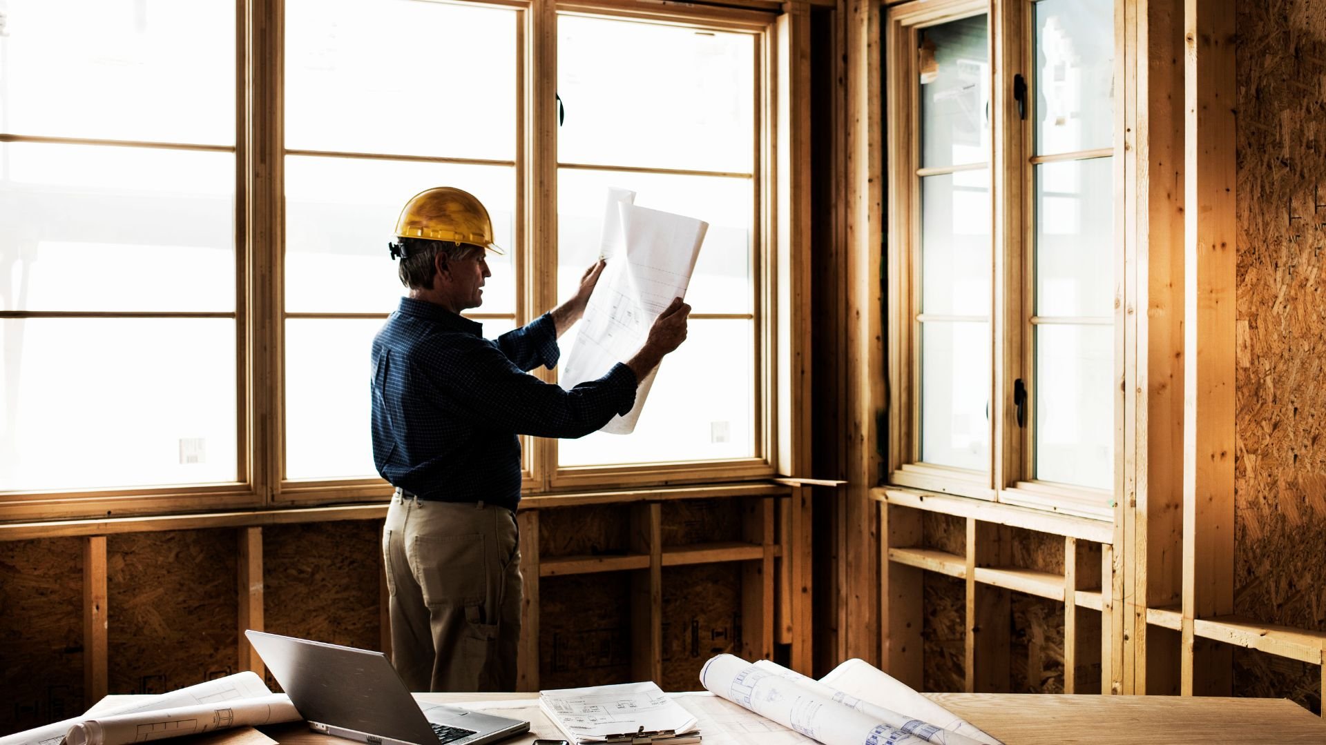 Construction worker reviewing blueprints in unfinished building with laptop